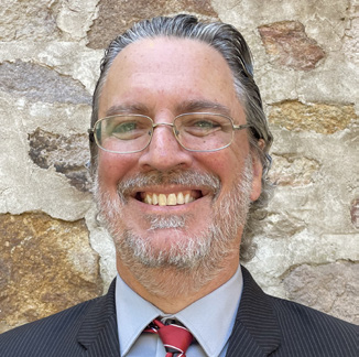Portrait of Kenneth Kinter with glasses, gray hair and beard wearing formal attire standing in front of a rock wall.